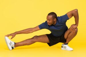 Sporty Black Male Doing Stetching Exercise Touching Toe, Yellow Background